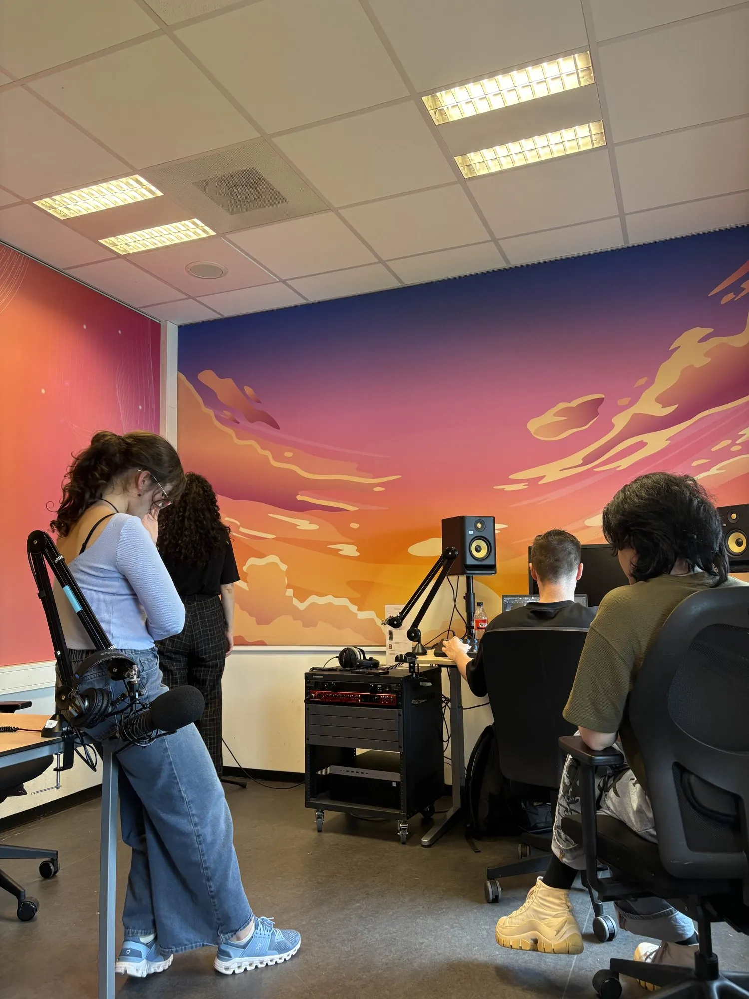 Team members recording vocals in a studio with a sunset mural on the wall